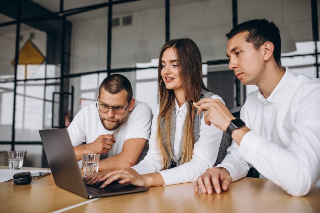 Three professionals working together at a table: a woman with long brown hair typing on a laptop, smiling, while two men in white shirts look at the screen — one with glasses and beard, the other holding a pen.