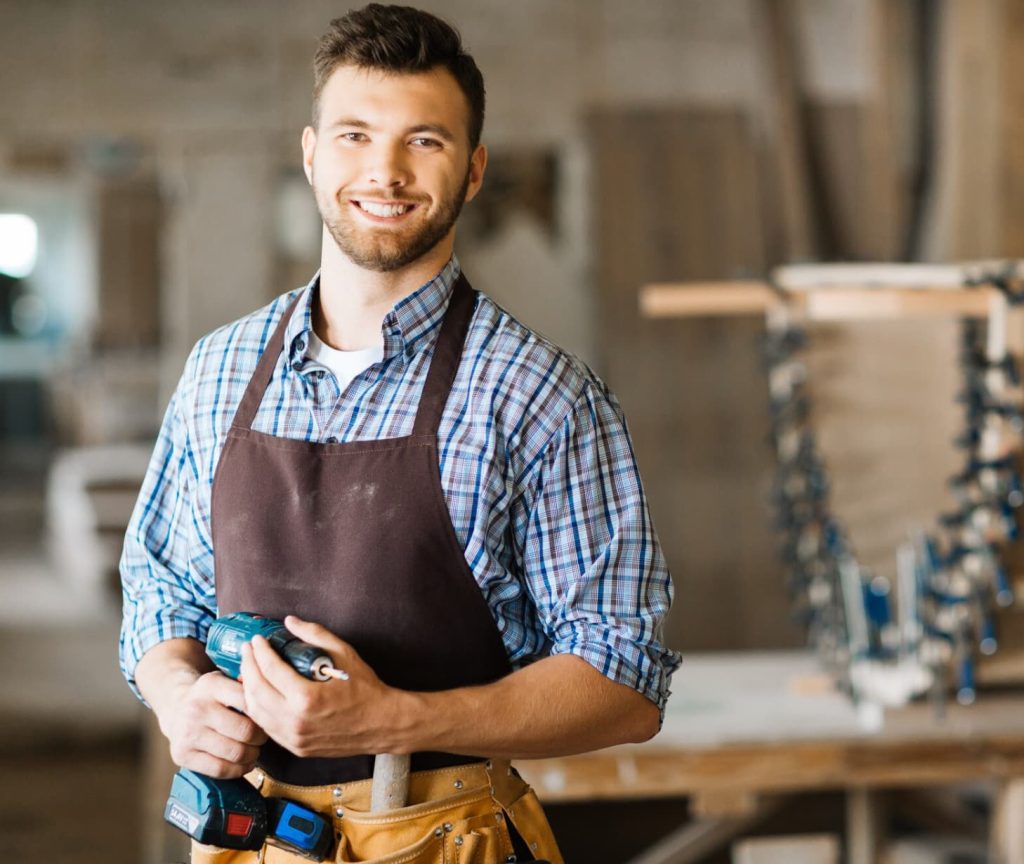 Smiling young carpenter with beard holding a power drill in a workshop.