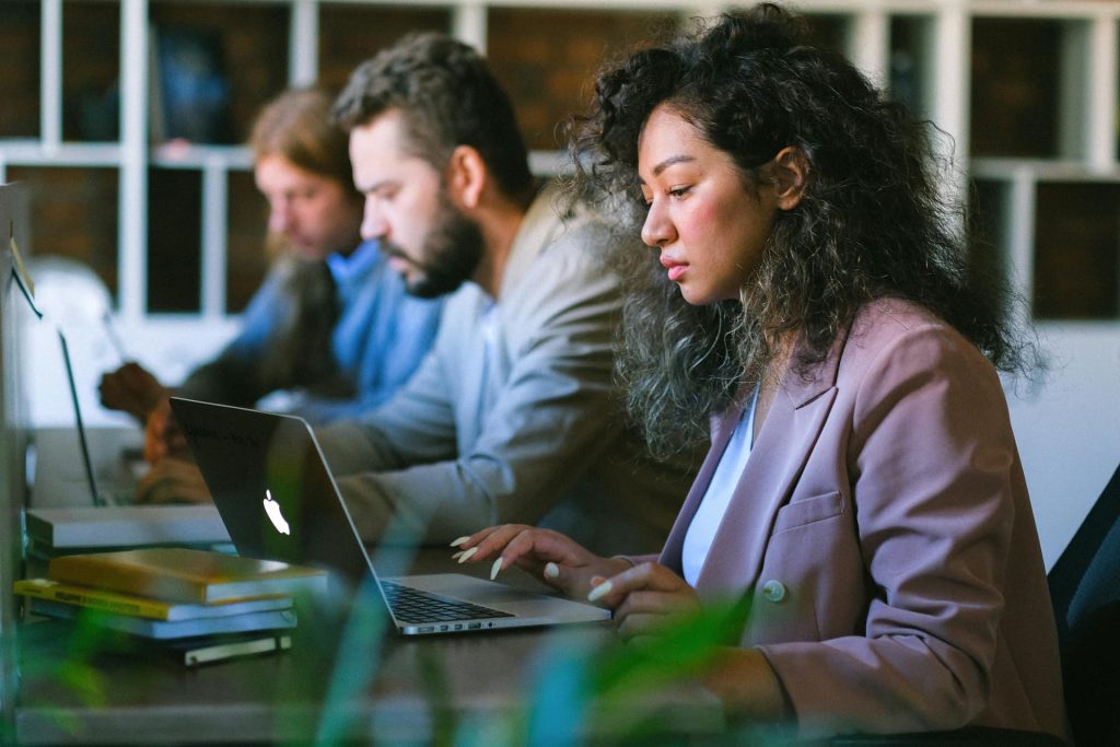 Woman with curly hair in a pink blazer typing on a MacBook in an office, with two men working in the background.